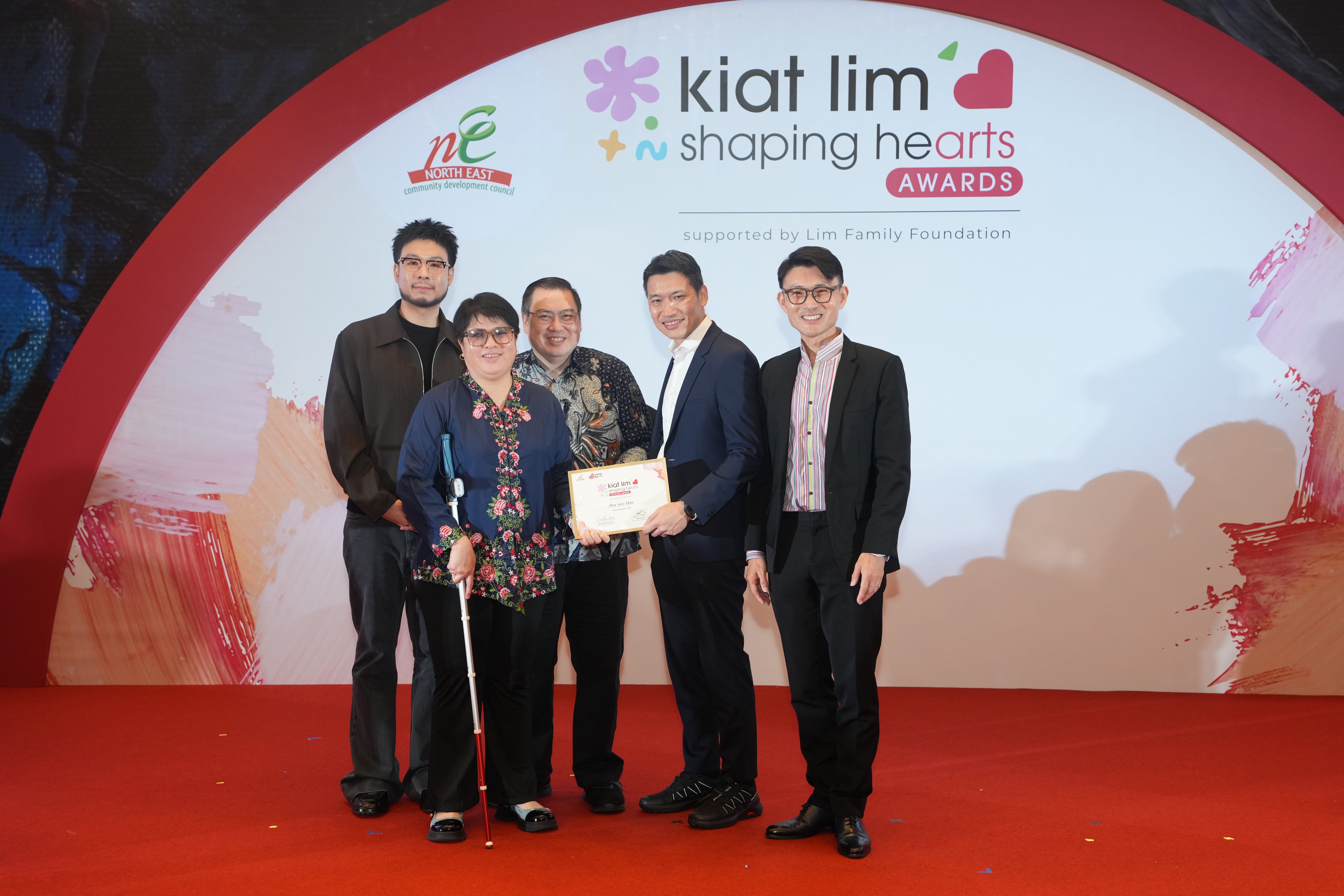 Award recipient standing on stage with the presenter, holding their certificate and posing for a group photo during the award ceremony, with a backdrop reading ‘Kiat Lim-Shaping Hearts Awards'.